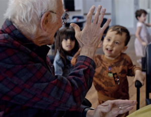 An older man sitting at a table offers a high five to an enthusiastic toddler.